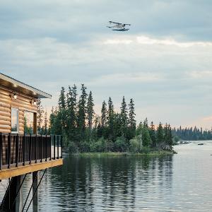 A floatplane flies over a calm lake towards a modern log cabin lodge at sunset in the Canadian wilderness.
