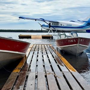 A blue and white Nakina Air Service floatplane docked behind two red aluminum fishing boats at a wooden pier on a calm lake.