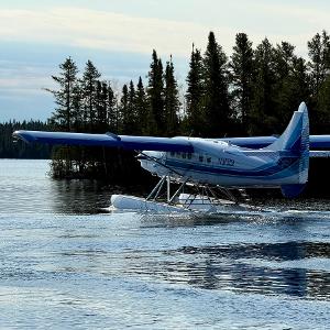 A white and blue Nakina Air Service floatplane taxiing on a calm lake surrounded by dense evergreen forest.