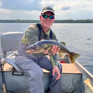 A fisherman in a boat holding a freshly caught walleye on a calm lake in the Canadian wilderness.