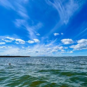 A vast blue lake under a dramatic sky with wispy white clouds and a distant treeline.